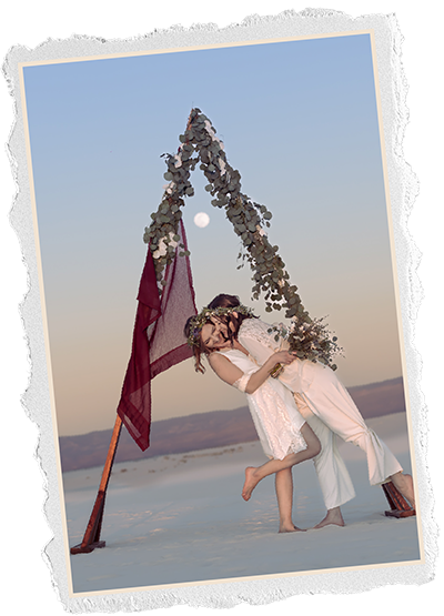 Torn edge photo of two brides embracing under a eucalyptus arch on white sand