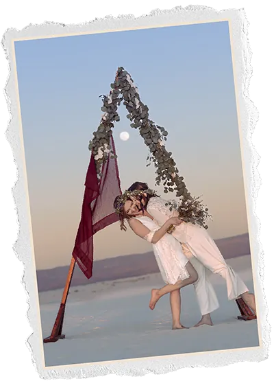 Torn edge photo of two brides embracing under a eucalyptus arch on white sand