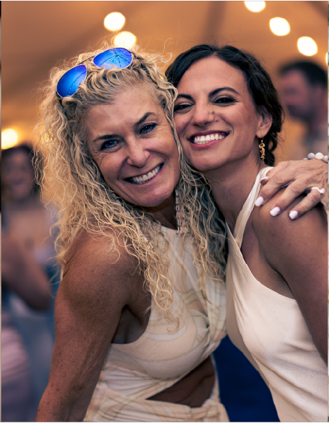 Two smiling women in cream dresses embrace under string lights