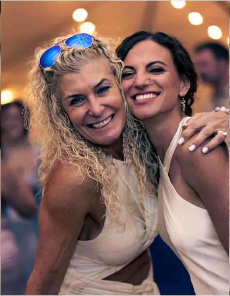 Two smiling women in cream dresses embrace under string lights