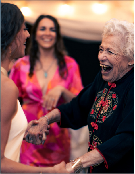 Laughing elderly woman holds hands with a woman in white at a party