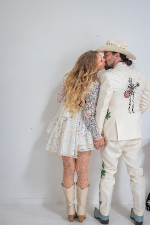 Mehama in a lace dress kisses her partner in a white western suit and cowboy hat