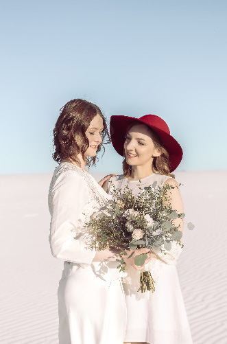 Two brides in white, one in a red hat, on white sand dunes with a bouquet