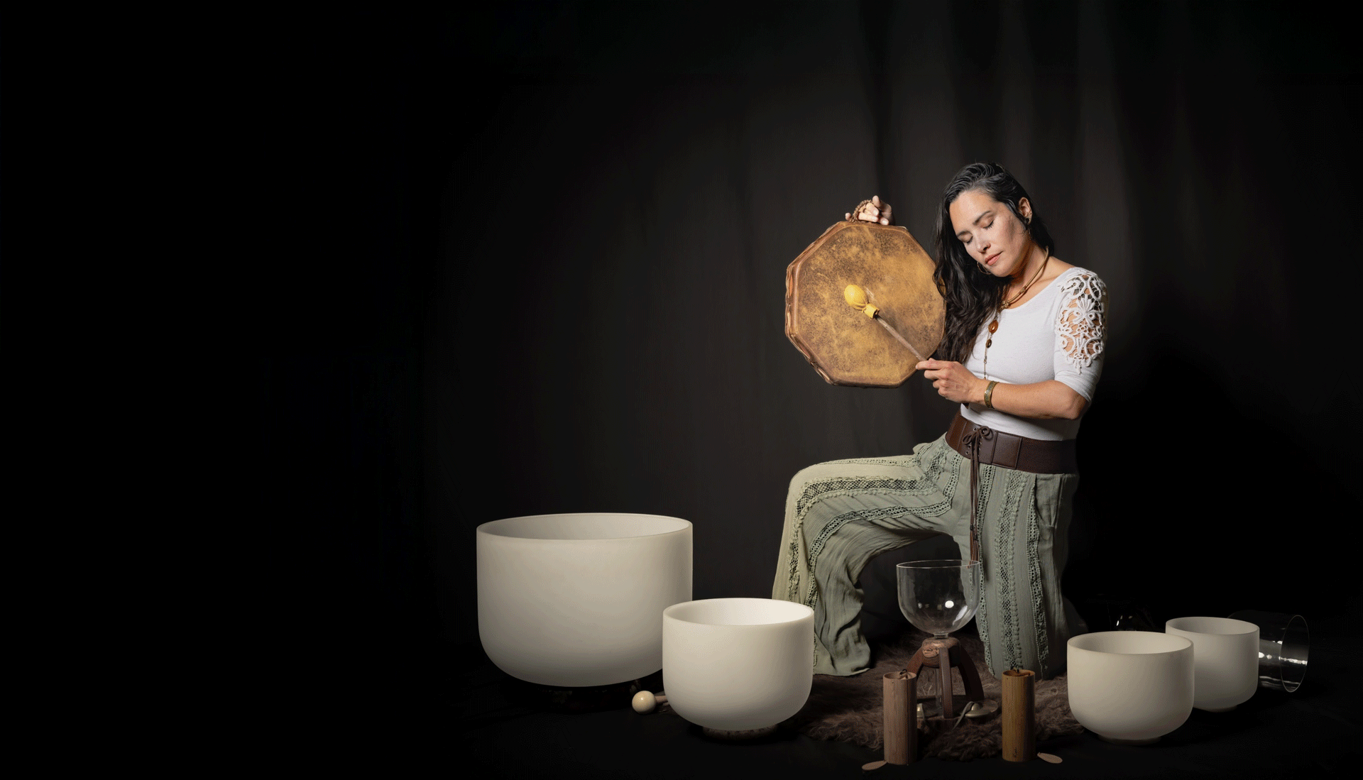 Sound healer plays a frame drum beside crystal singing bowls on a dark set