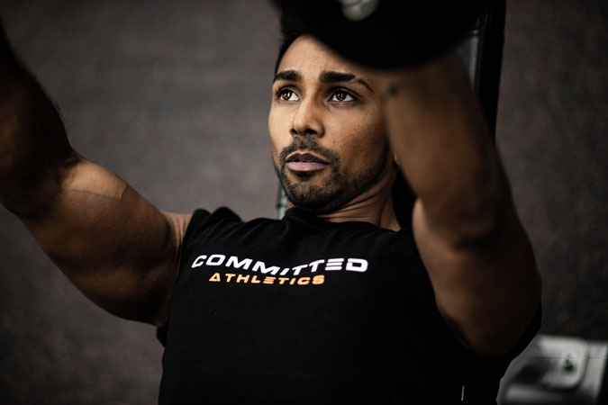 Athlete in a Committed Athletics shirt lifts weights on a gym bench