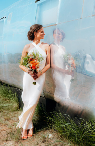 Bride with an orange bouquet leans against a reflective silver trailer
