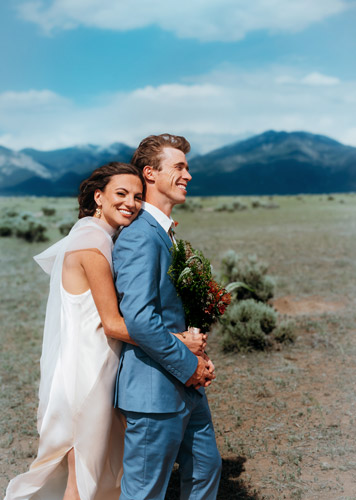 Bride hugs groom in a blue suit in a sagebrush field below mountains