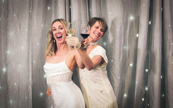 Two women in white raise wine glasses in front of a twinkling curtain