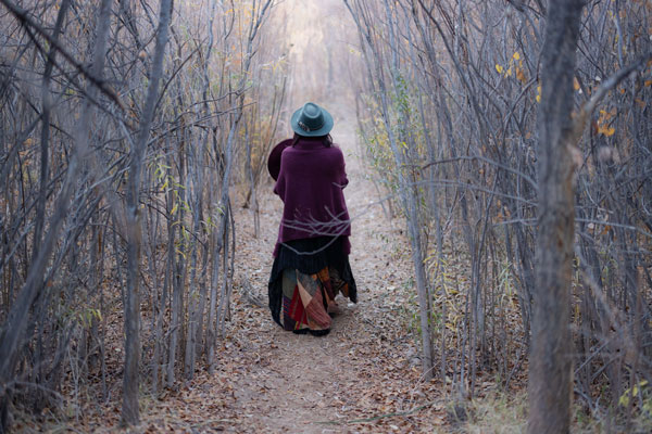 Woman in a wide brim hat and patchwork skirt walks a wooded autumn path