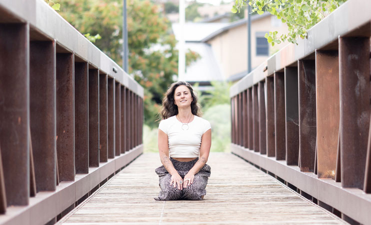 Yoga instructor kneels on a wooden footbridge smiling at the camera