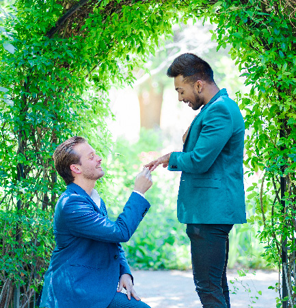 Man kneels to propose to his partner under a green leafy arch