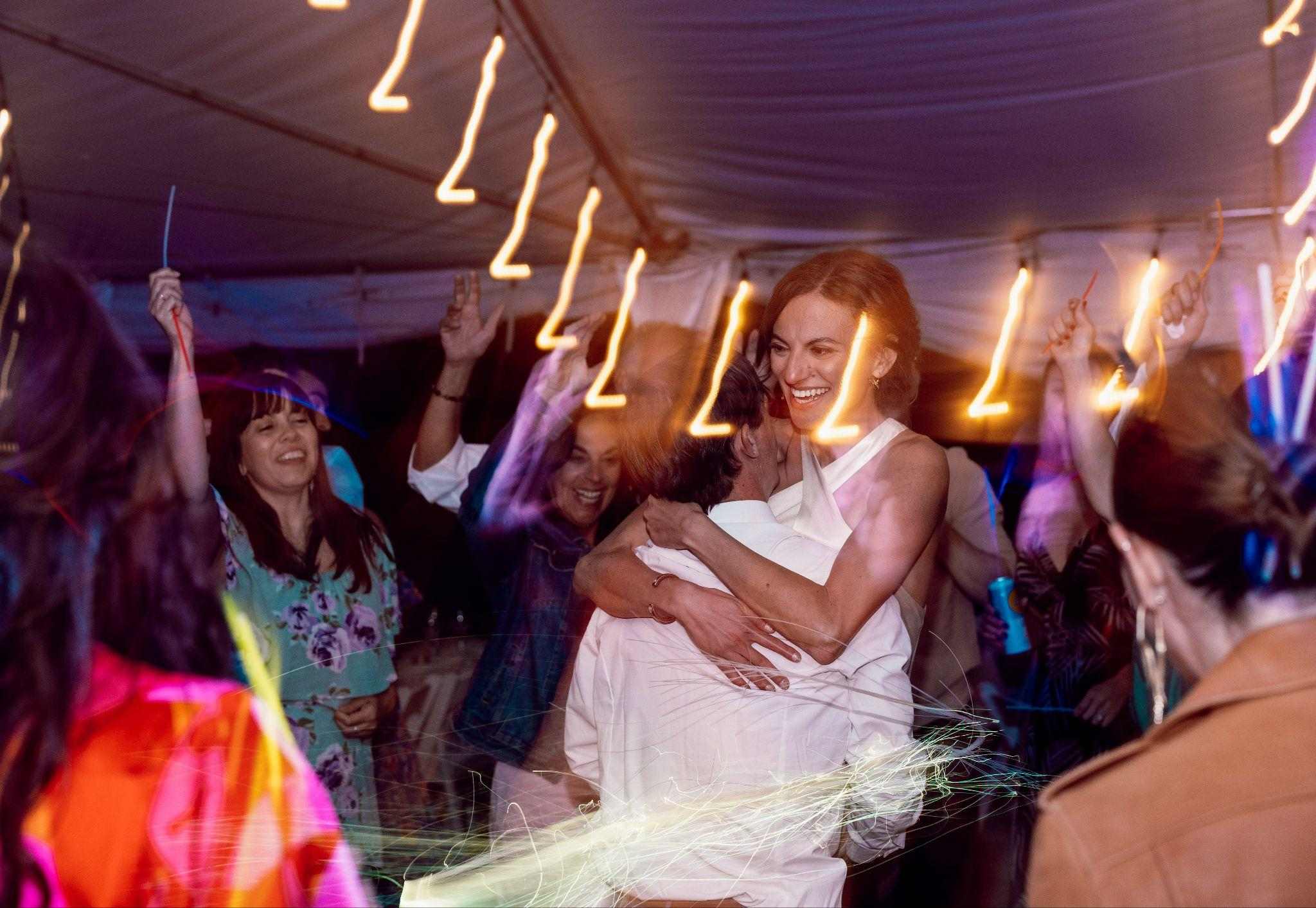 Bride hugs a guest on a packed dance floor with light trails overhead