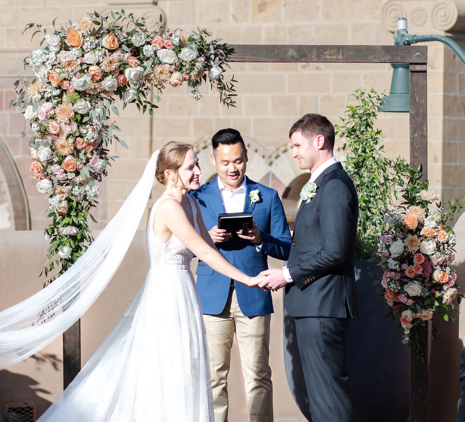 Bride and groom holding hands with the officiant under a floral arch at an adobe venue
