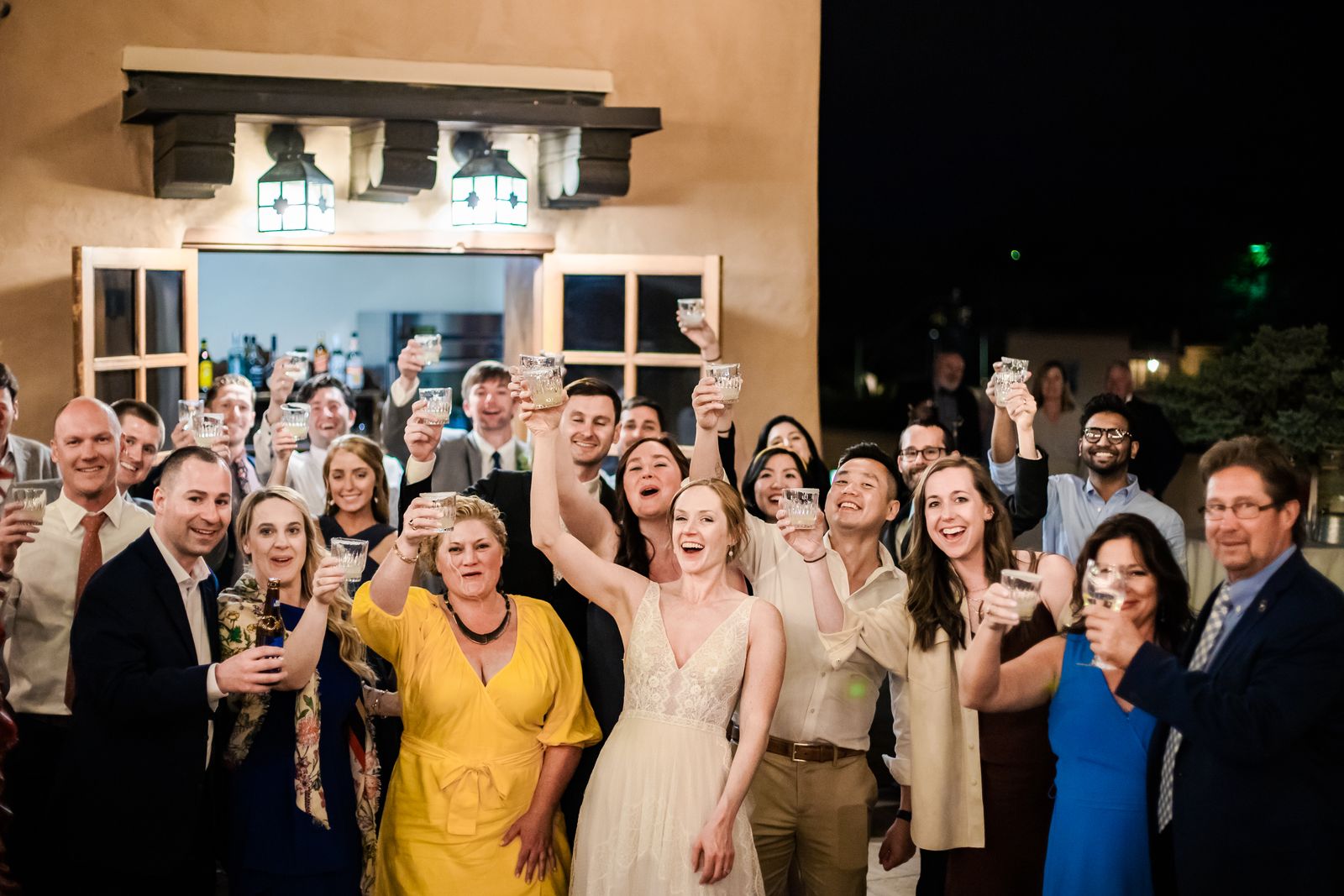 Bride and guests cheering with raised glasses outside an adobe building at night