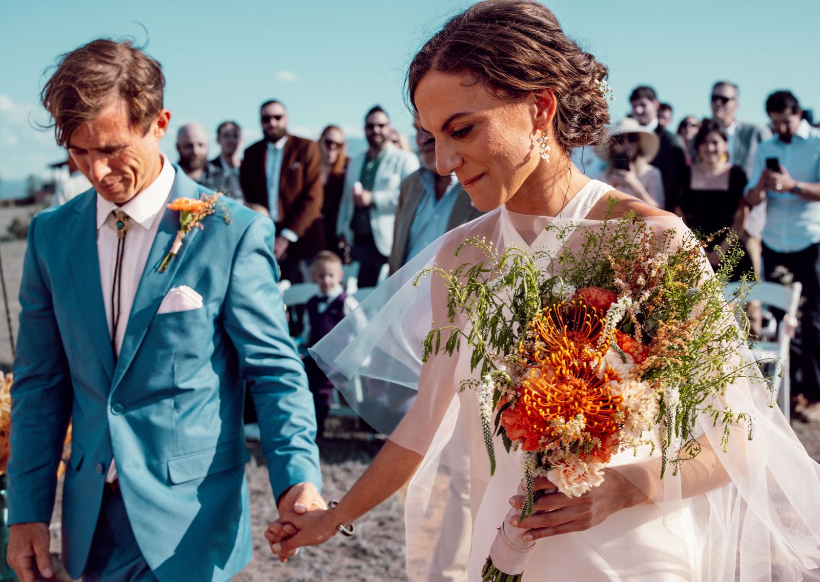 Groom in a teal suit and bride with an orange bouquet walking hand in hand past guests