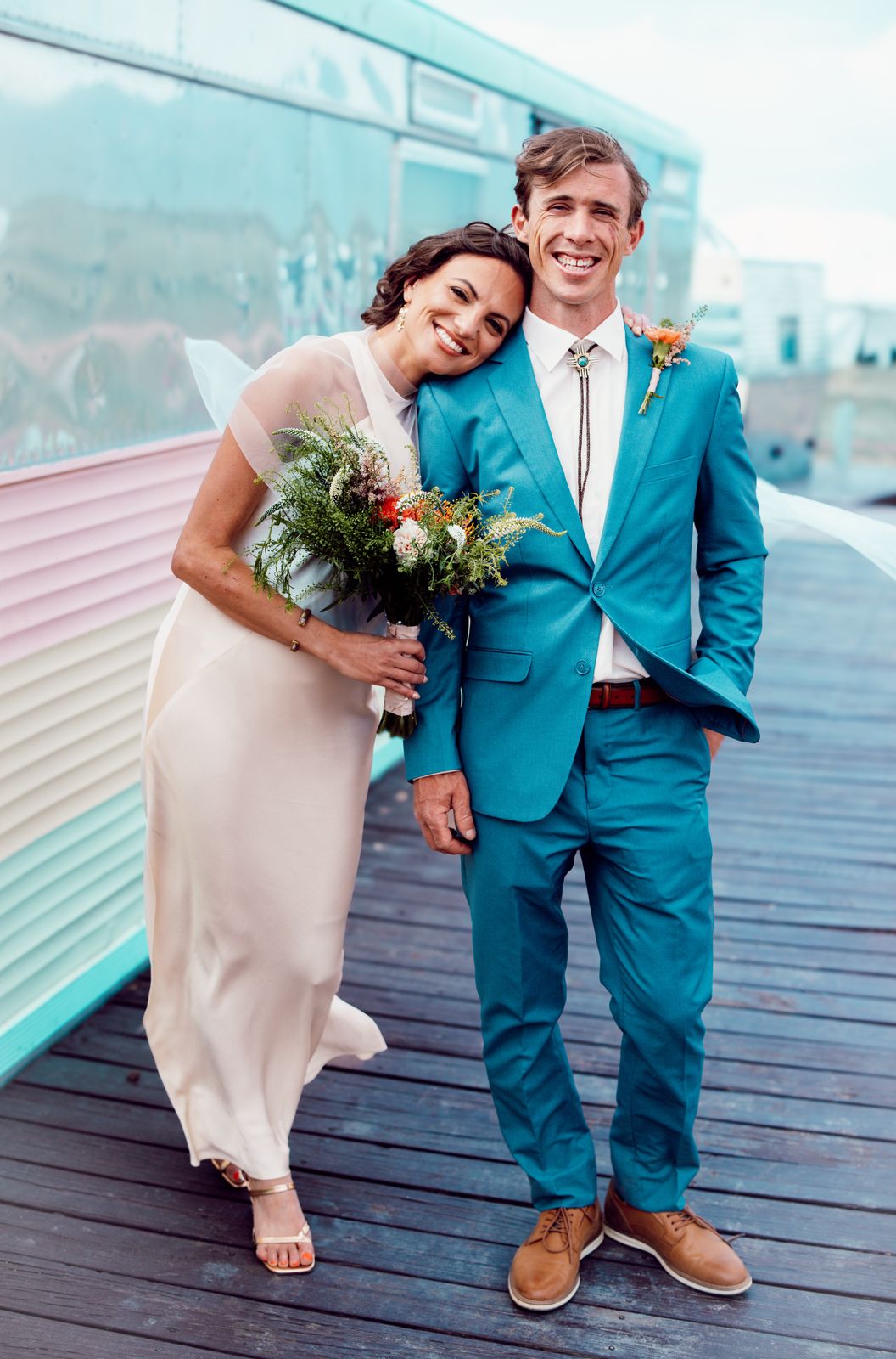 Bride and groom in a teal suit smiling on a boardwalk next to a pastel trailer
