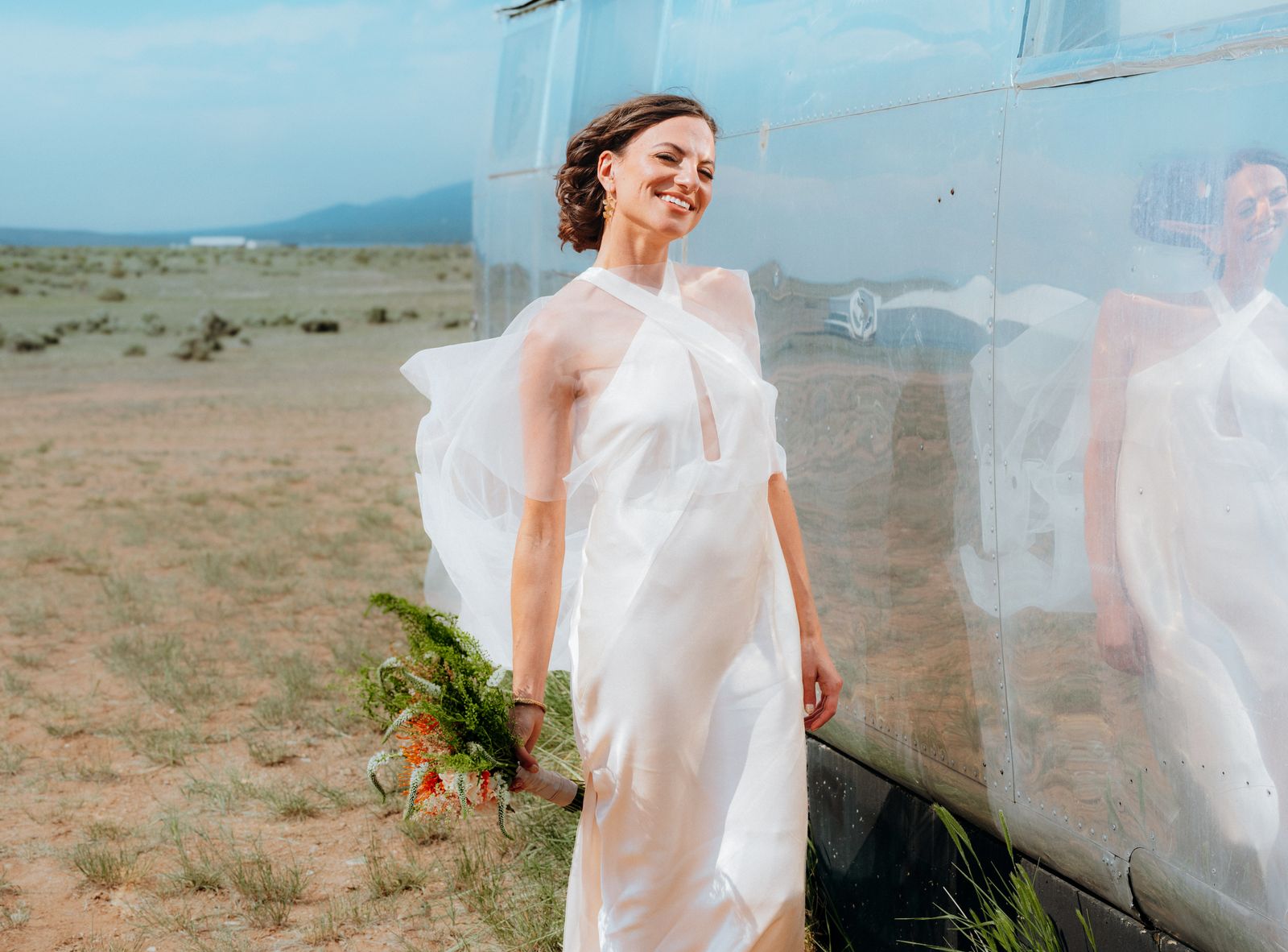 Bride in a sheer gown leaning against a silver trailer in the desert