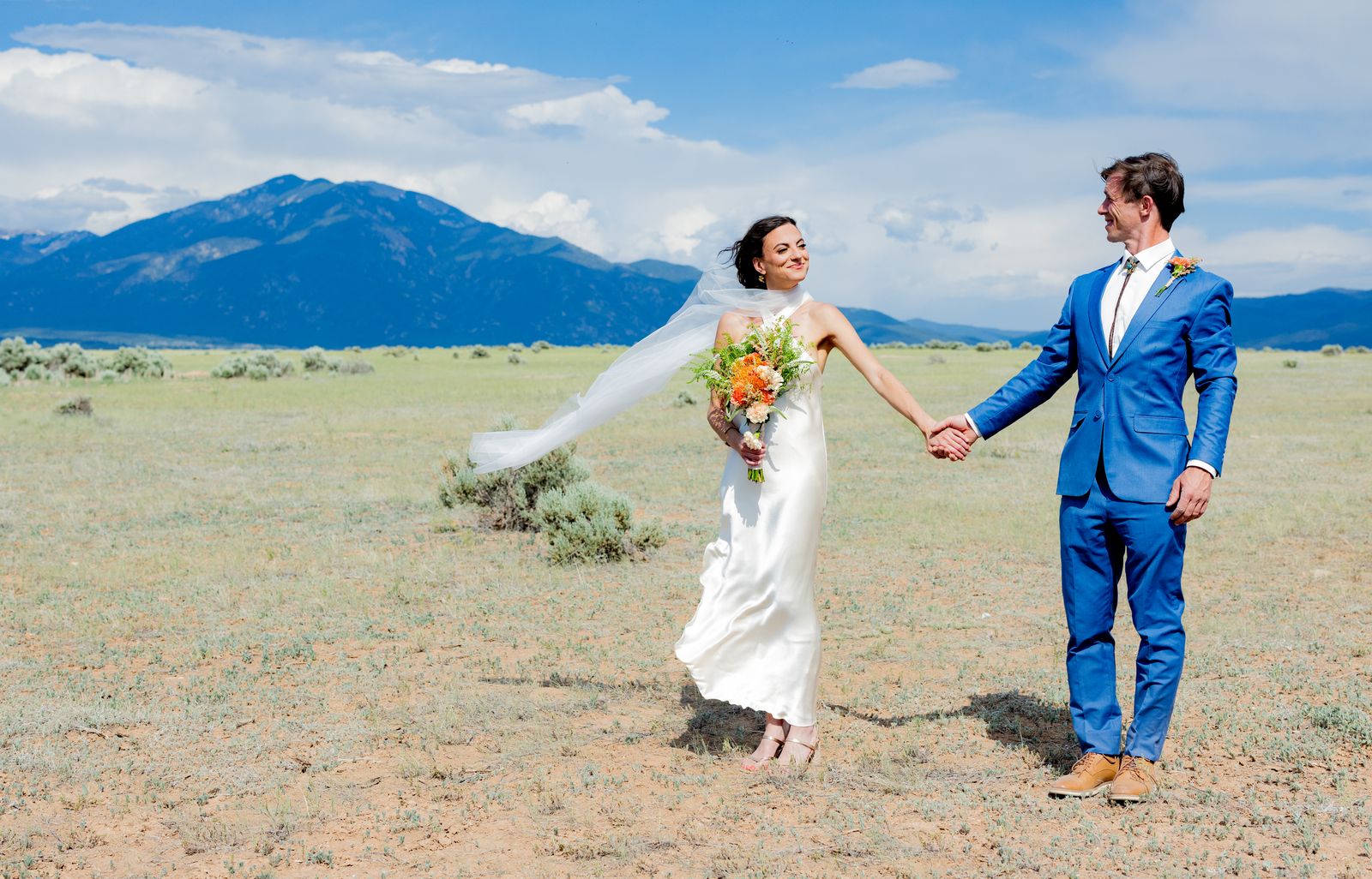 Bride with her veil flying holding the groom's hand in a field below the mountains