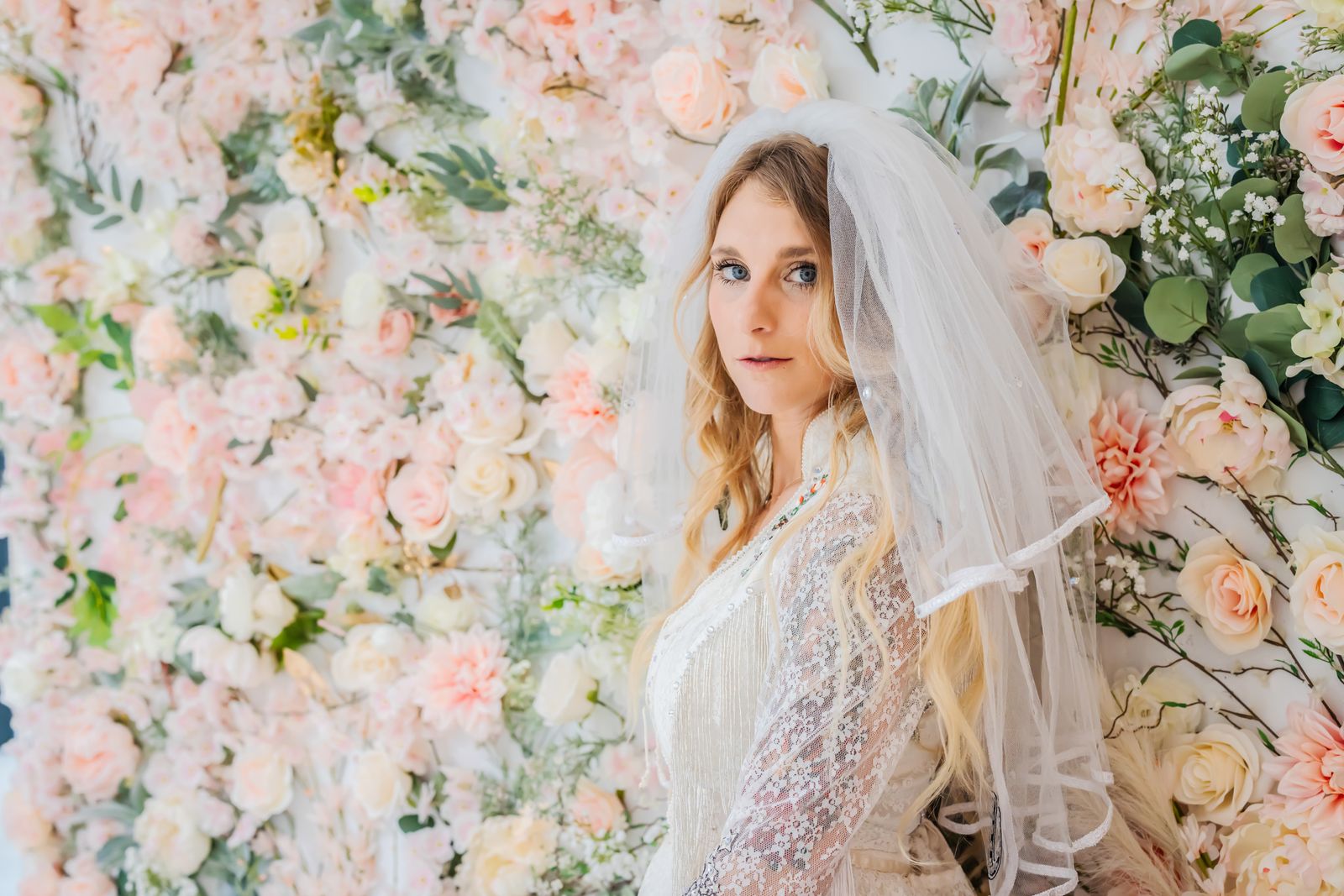 Bride in a veil and lace jacket posing in front of a flower wall