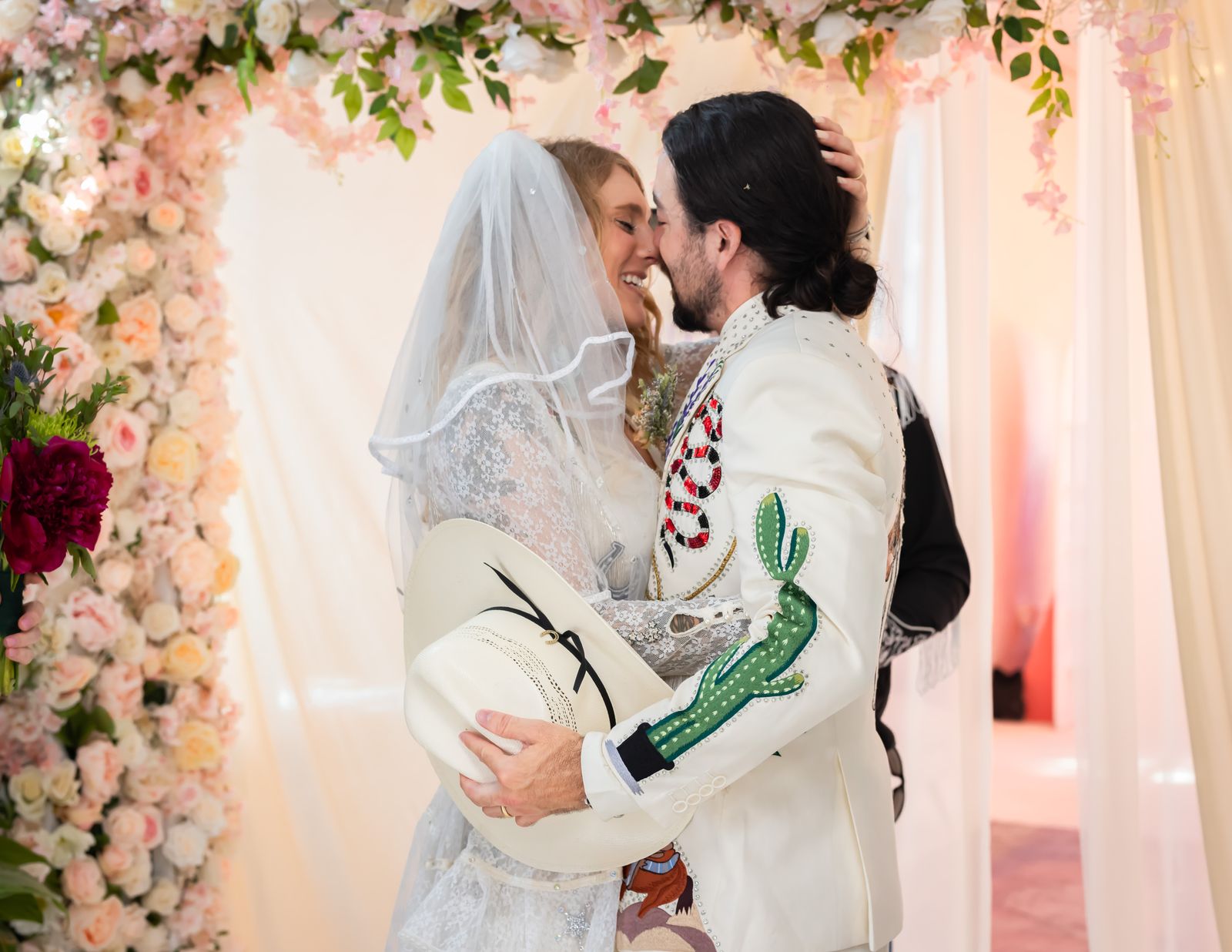 Groom kissing the bride under a floral arch while holding his cowboy hat