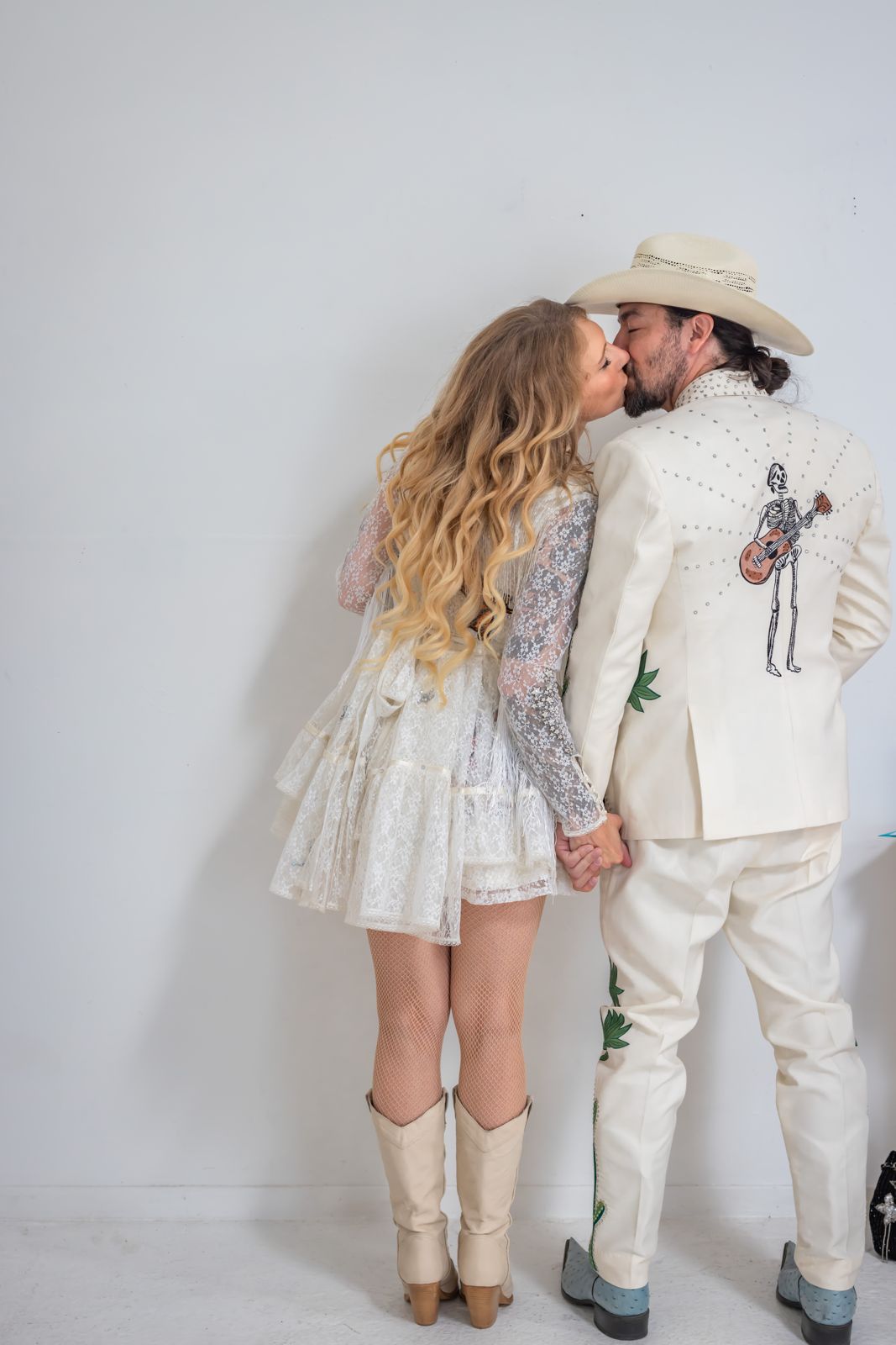 Couple in white western outfits kissing and holding hands against a white wall