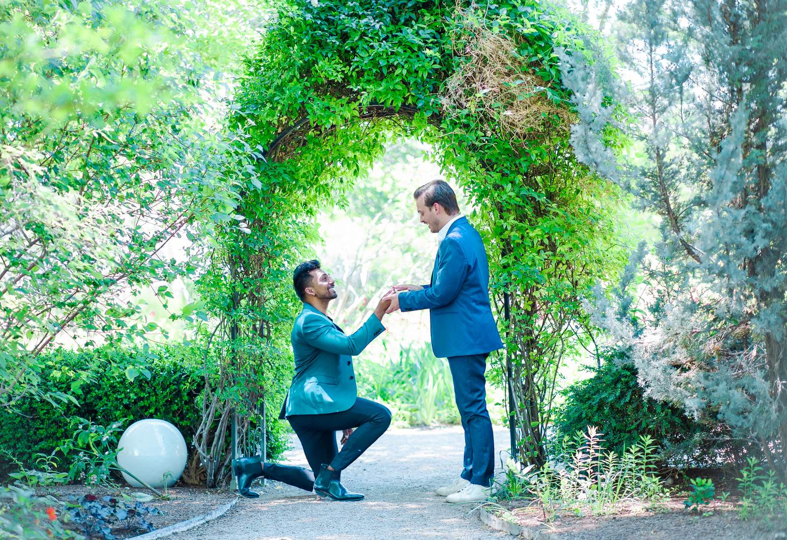 Groom kneeling to propose to his partner beneath a leafy green arch