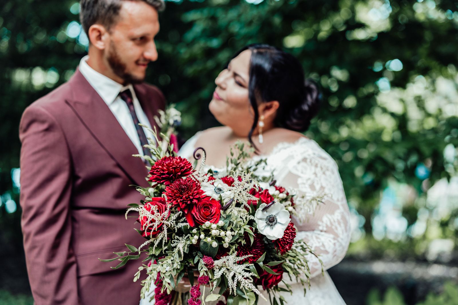 Groom in a burgundy suit and bride with a red bouquet gazing at each other