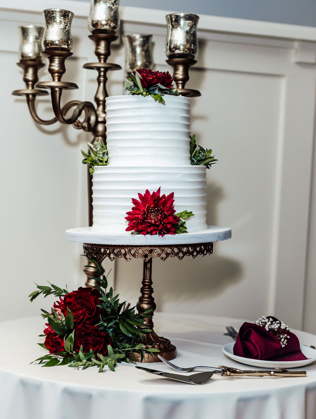 Three tier white cake with red flowers beneath a brass candelabra
