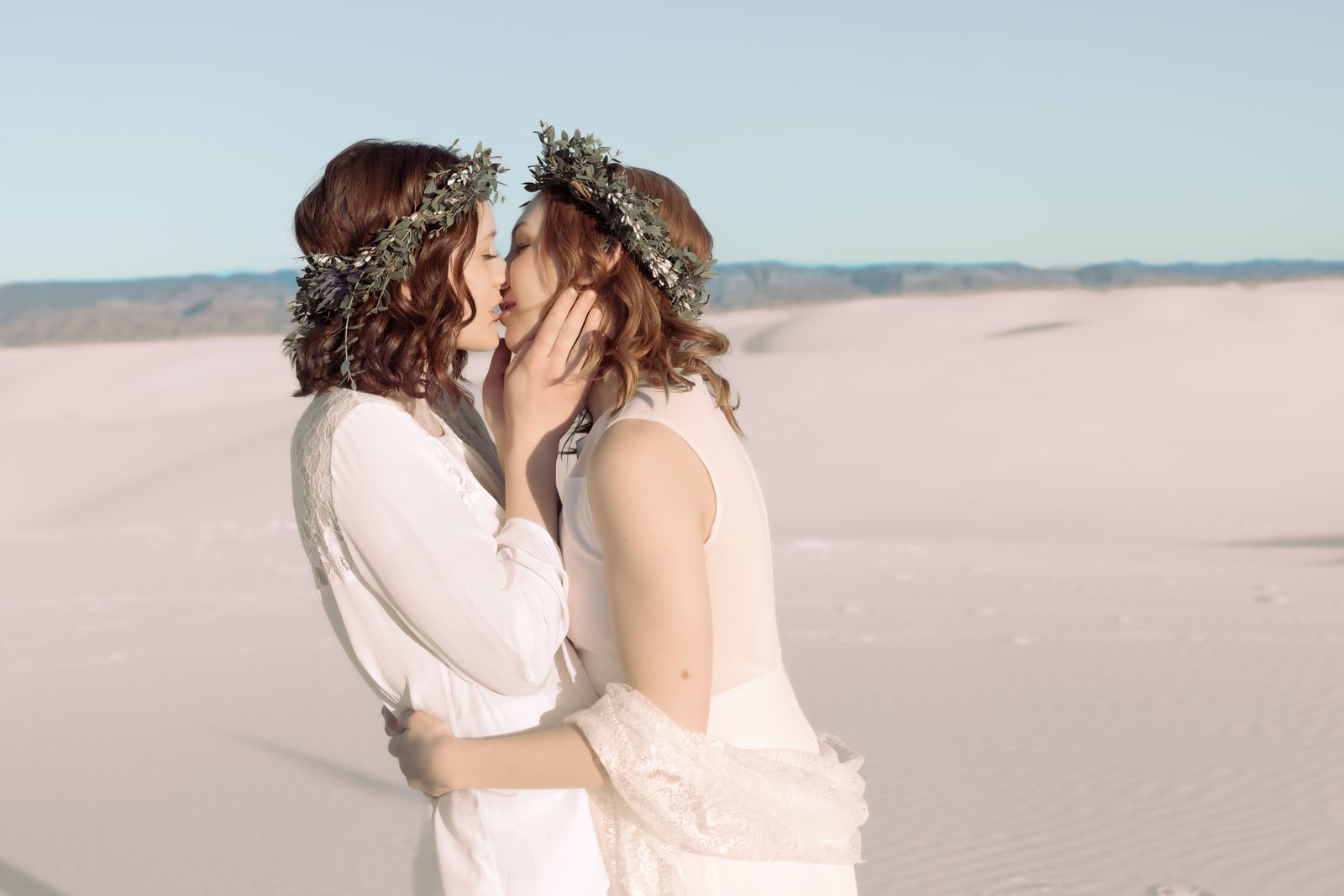 Two brides in flower crowns kissing on white sand dunes