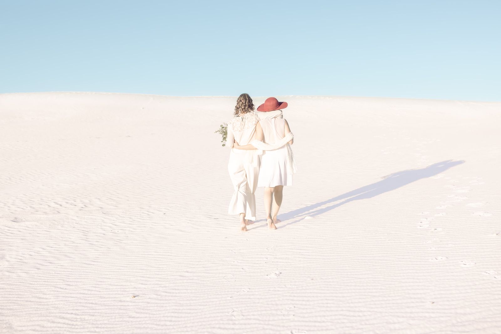 Two brides walking arm in arm across white sand, one in a red hat