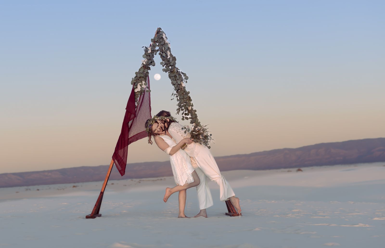 Two brides embracing under a triangular floral arch on the dunes at dusk