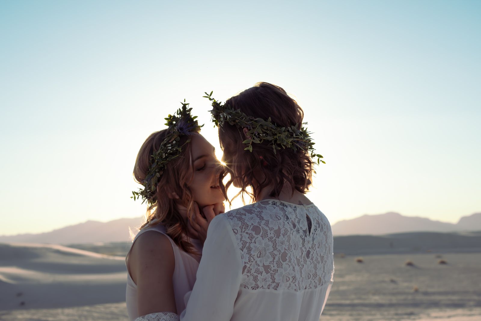 Two brides in flower crowns touching foreheads against a bright sky