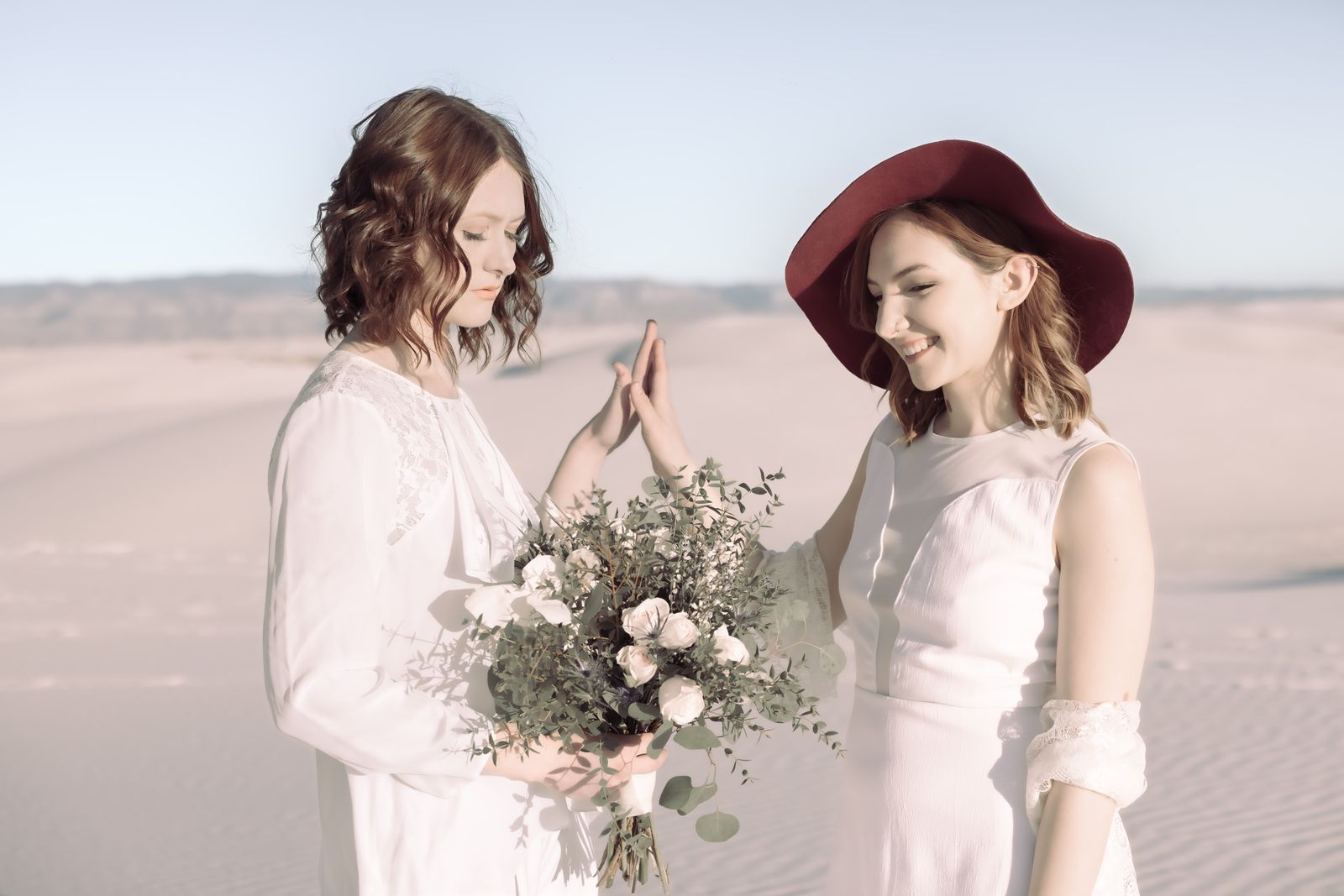Two brides smiling and holding hands, one in a wide red hat, with a bouquet