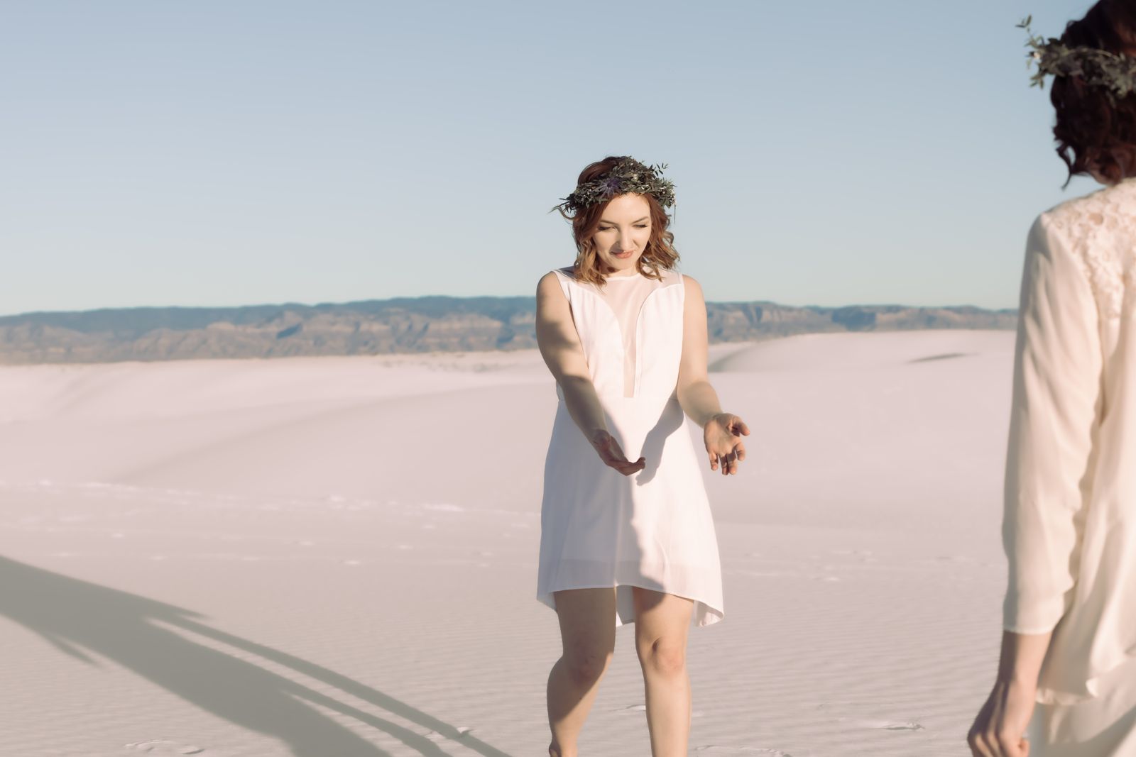 Bride in a flower crown smiling and reaching toward her partner on the dunes