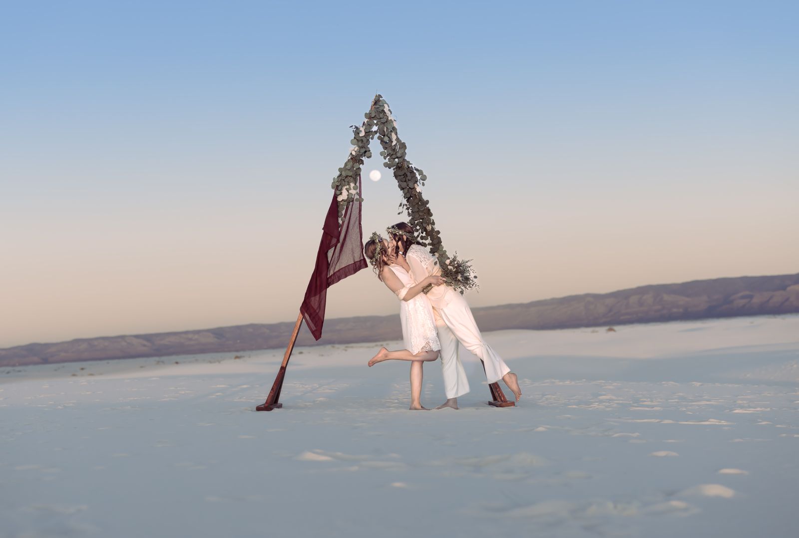 Two brides kissing under a triangular floral arch at dusk