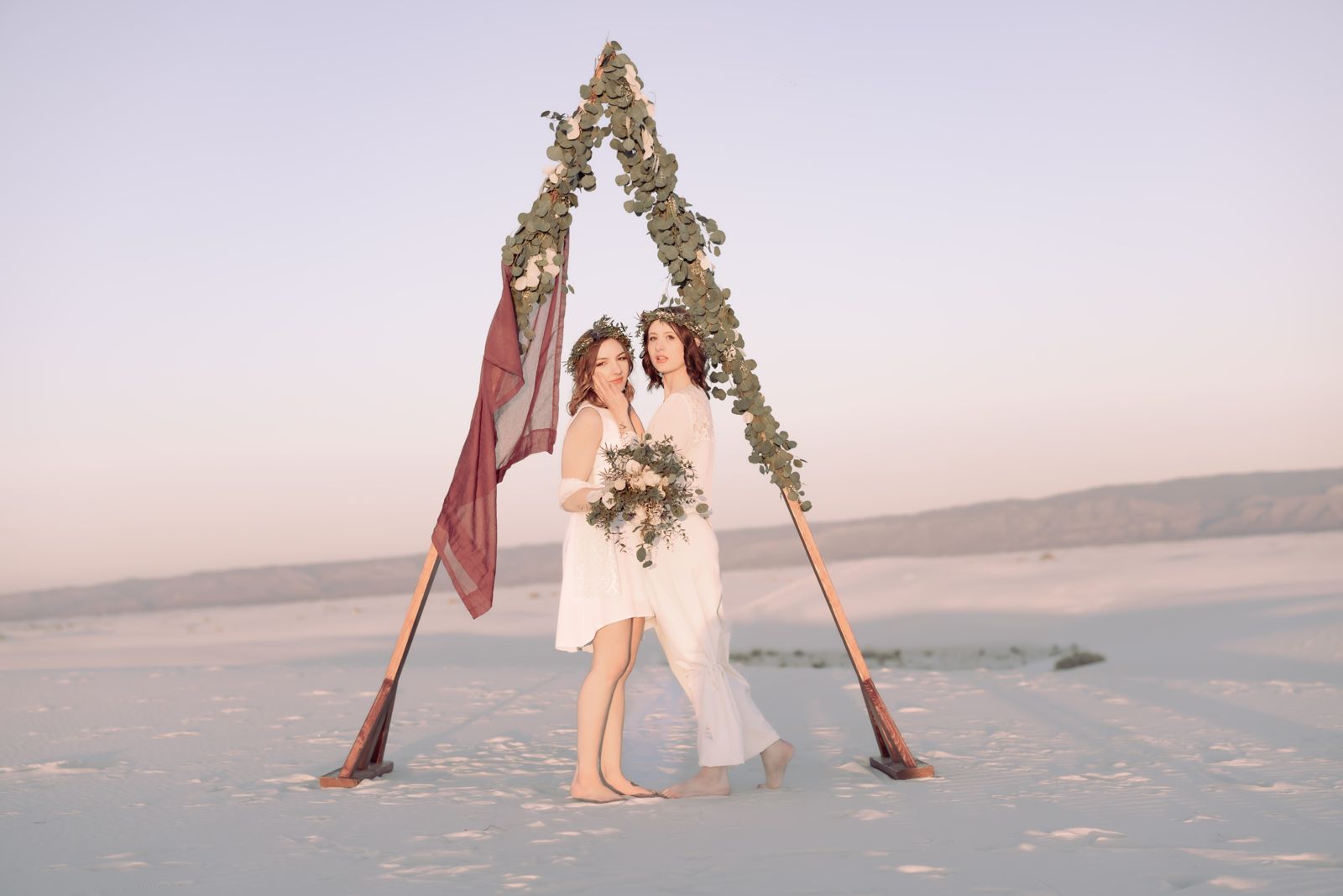 Two brides laughing beneath a floral arch, one in a red hat, with the moon rising